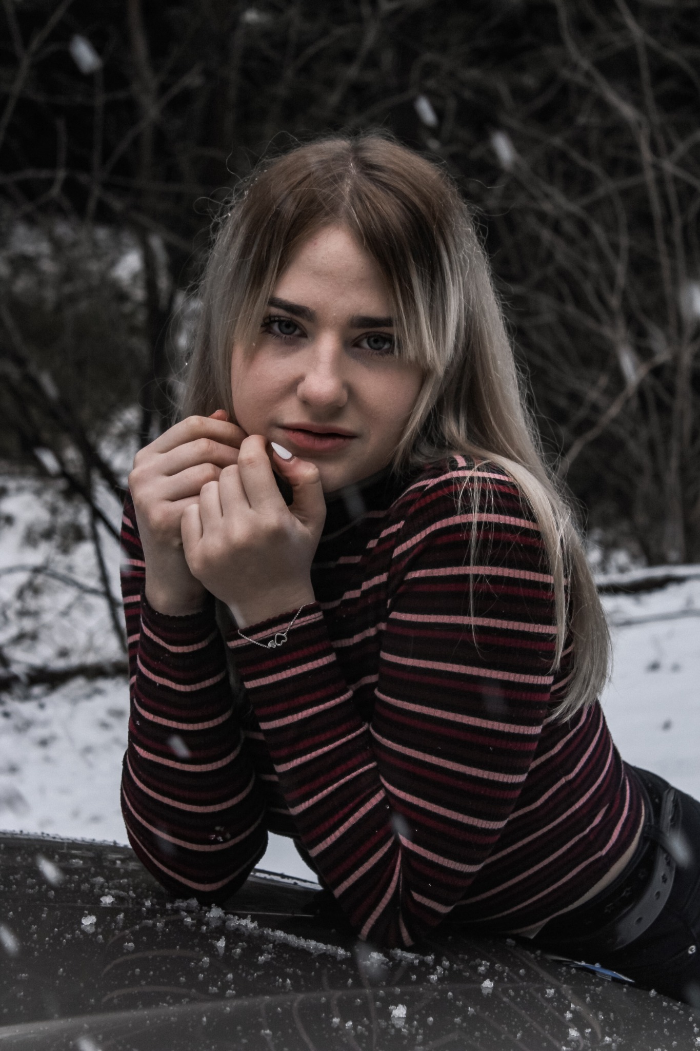 Młoda kobieta z blond włosami w bluzce w paski pozuje w zimowej scenerii, opierając się o ciemną powierzchnię, z widoczną srebrną bransoletką na nadgarstku i padającym śniegiem w tle.