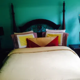 Ustawione łóżko z czterema słupkami, jasną pościelą i dekoracyjnymi poduszkami na tle turkusowej ściany, z lampą na szafce nocnej po lewej i wazonem z suchymi kwiatami po prawej.