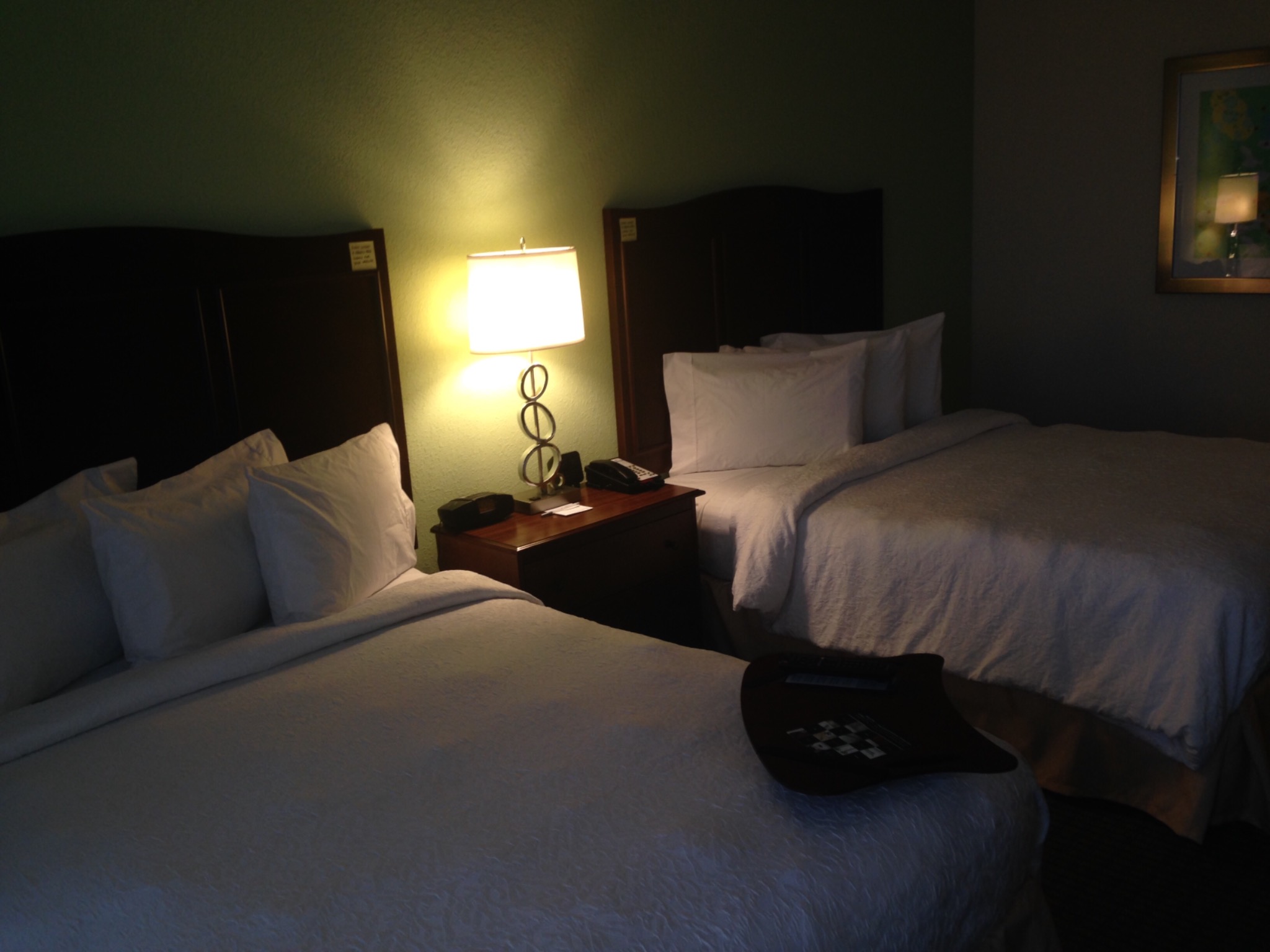 Dwa łóżka pojedyncze z białymi poduszkami i kołdrami w pokoju hotelowym, na jednym łóżku taca z szachownicą, stolik nocny z lampką i telefonem, obraz na ścianie.