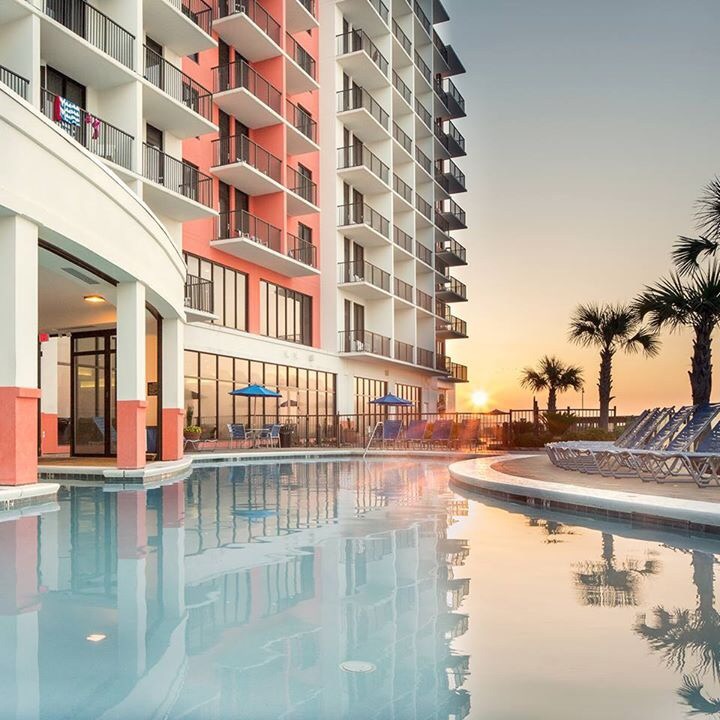 Wysoki budynek z balkonami w kolorach białym i łososiowym, odbijający się w basenie o zachodzie słońca, z palmami w tle.