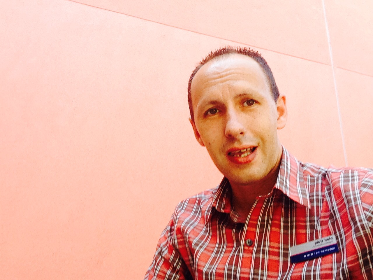 Portret mężczyzny w czerwonej koszuli w kratę z identyfikatorem, na tle jasnoróżowej ściany.