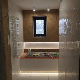 Bartosz Rosłan - Łazienka w trakcie wykańczania: widoczne płytki imitujące drewno na ścianie, białe płytki z geometrycznym wzorem, grzejnik drabinkowy, okno, podświetlenie LED pod półką i narzędzia glazurnicze...