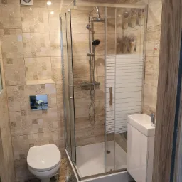 Nowoczesna łazienka z kabiną prysznicową, umywalką nablatową na szafce, toaletą i beżowymi płytkami z dekoracyjnym wzorem, wentylator w rogu pod sufitem.