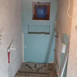 Wnętrze łazienki w trakcie prac: widoczne rury ogrzewania podłogowego, ściany częściowo pokryte płytami gipsowo-kartonowymi w kolorze turkusowym, okno zabezpieczone folią, instalacja...