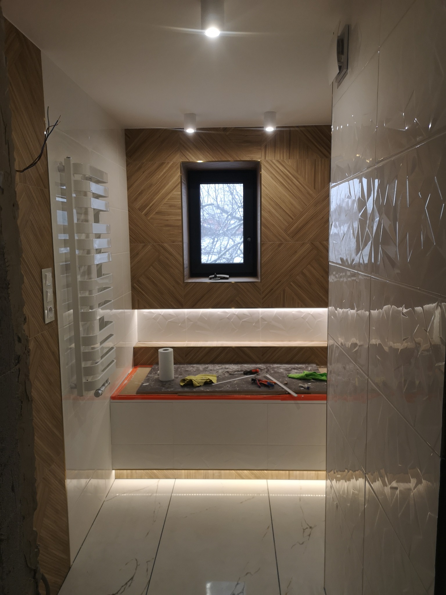 Łazienka w trakcie wykańczania: widoczne płytki imitujące drewno na ścianie, białe płytki z geometrycznym wzorem, grzejnik drabinkowy, okno, podświetlenie LED pod półką i narzędzia glazurnicze...