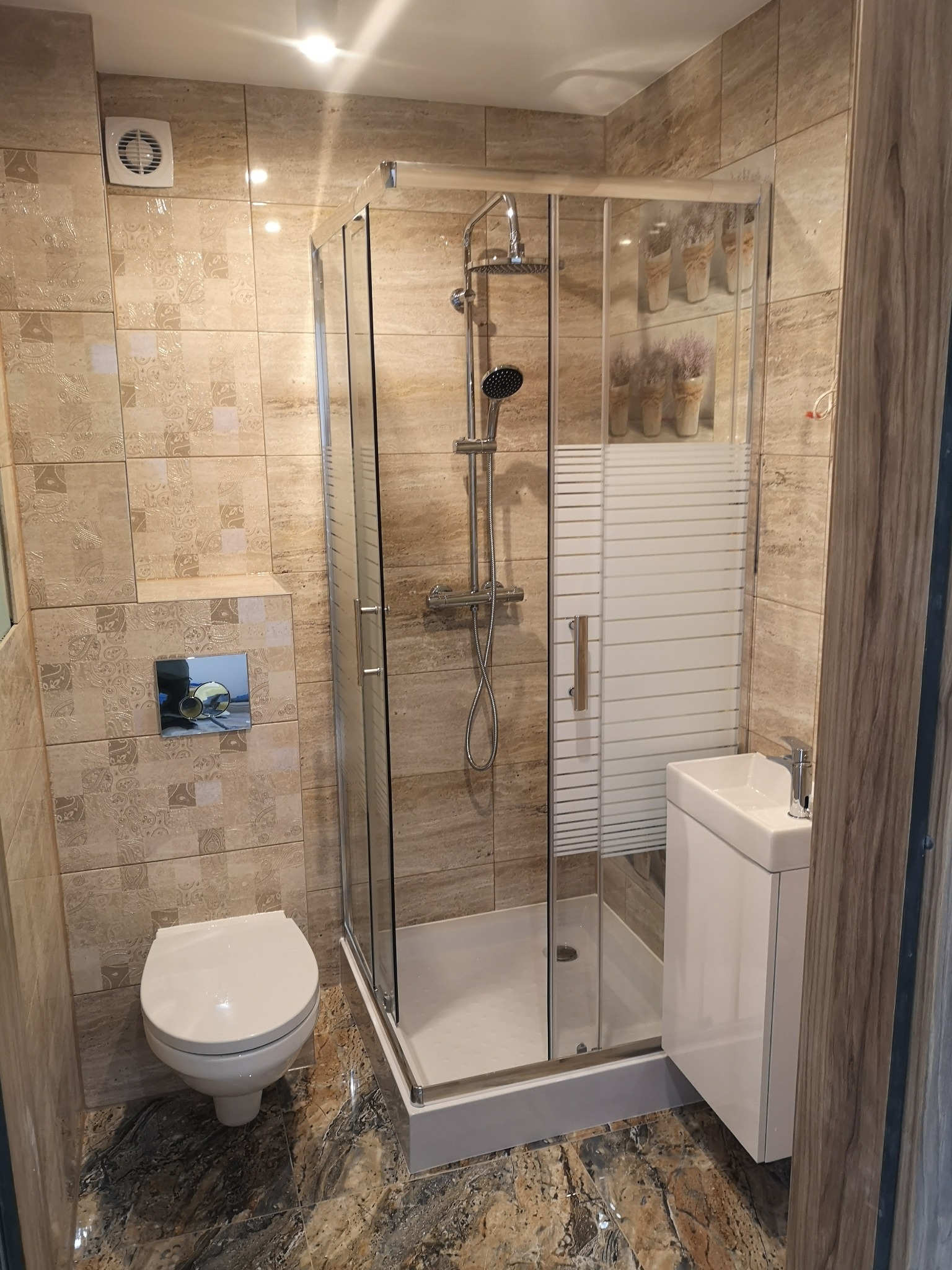 Nowoczesna łazienka z kabiną prysznicową, umywalką nablatową na szafce, toaletą i beżowymi płytkami z dekoracyjnym wzorem, wentylator w rogu pod sufitem.
