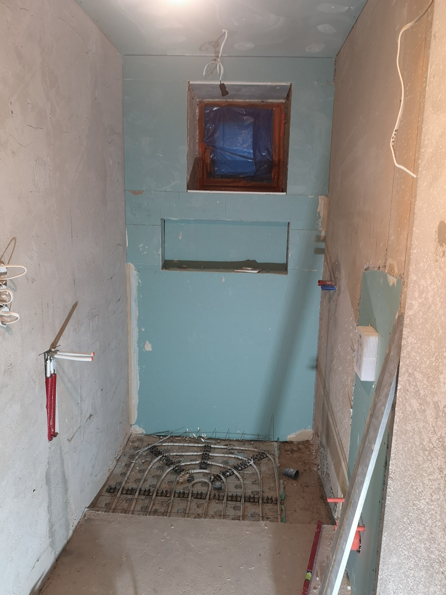 Wnętrze łazienki w trakcie prac: widoczne rury ogrzewania podłogowego, ściany częściowo pokryte płytami gipsowo-kartonowymi w kolorze turkusowym, okno zabezpieczone folią, instalacja...