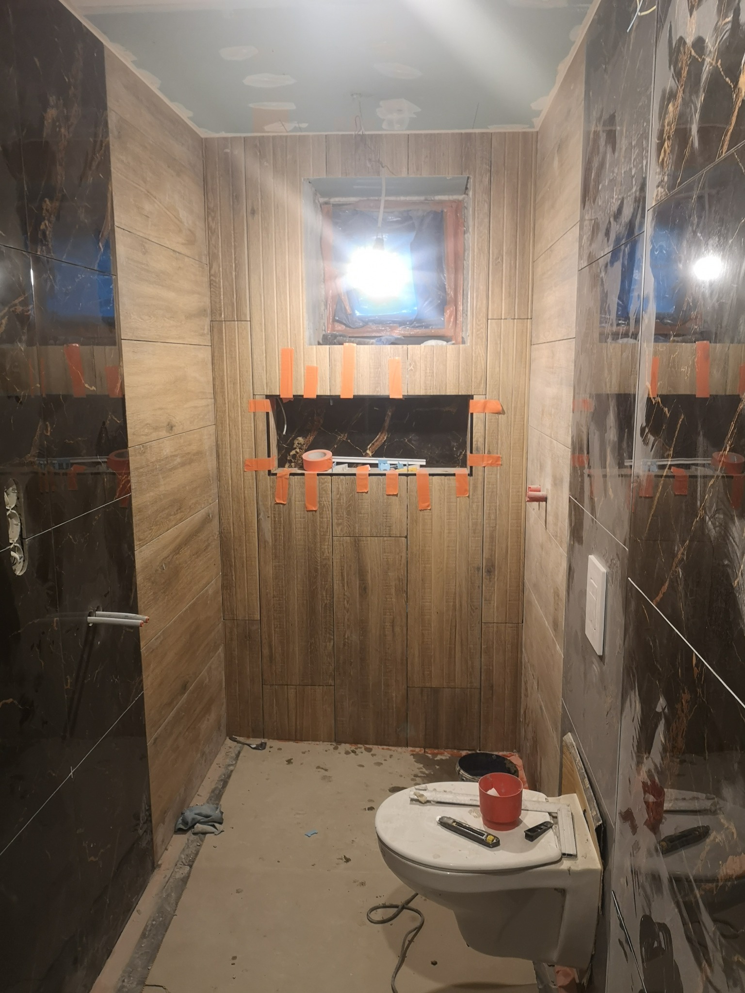 Łazienka w trakcie remontu: brązowe i czarne płytki, widoczne narzędzia na sedesie, pomarańczowa taśma zabezpieczająca płytki, surowy sufit.