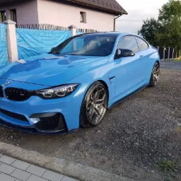 Błękitne BMW M4 po renowacji lakieru, zaparkowane na żwirowym podjeździe, widoczne detale: czarne felgi i karbonowy splitter.