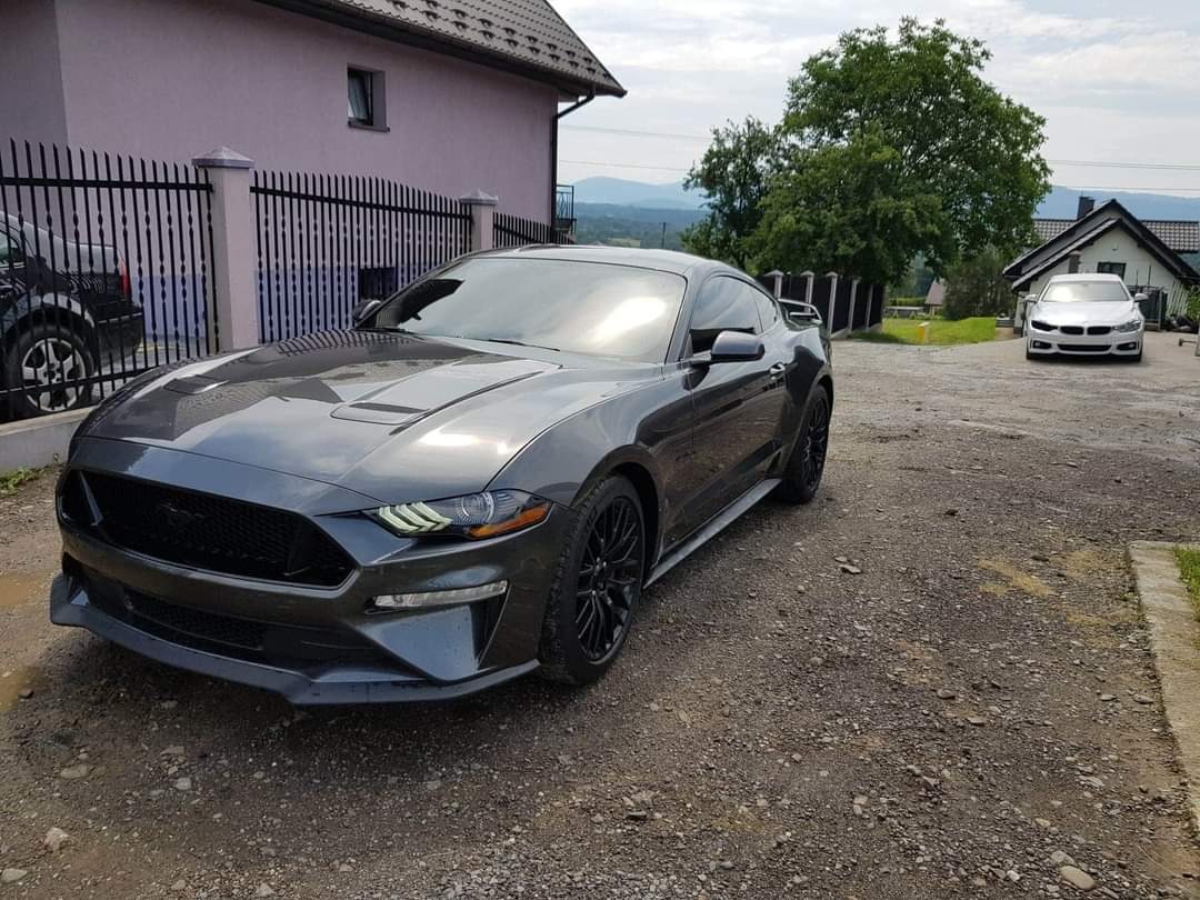 Szary Ford Mustang zaparkowany na żwirowym podjeździe przed domem z czarnym ogrodzeniem, w tle widoczny drugi samochód i zalesione wzgórza.