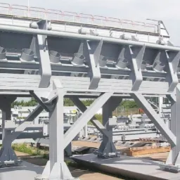 Szara, stalowa konstrukcja nośna z rzędami okrągłych otworów, ustawiona na zewnątrz w słoneczny dzień, z elementami rusztowania i zielenią w tle.