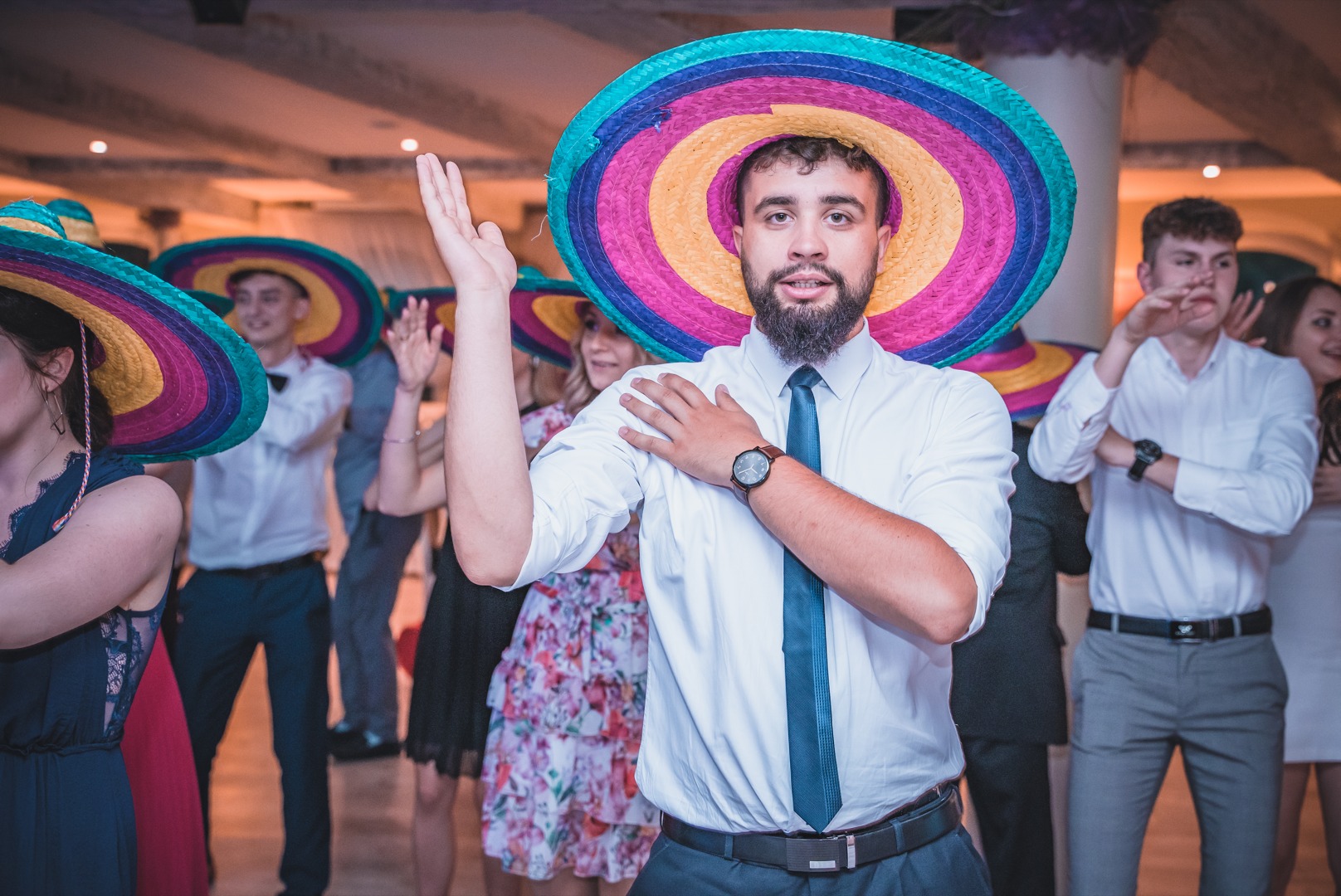 Mężczyzna w białej koszuli i krawacie, z brodą, noszący kolorowe sombrero na tle bawiących się osób, prawdopodobnie na imprezie.