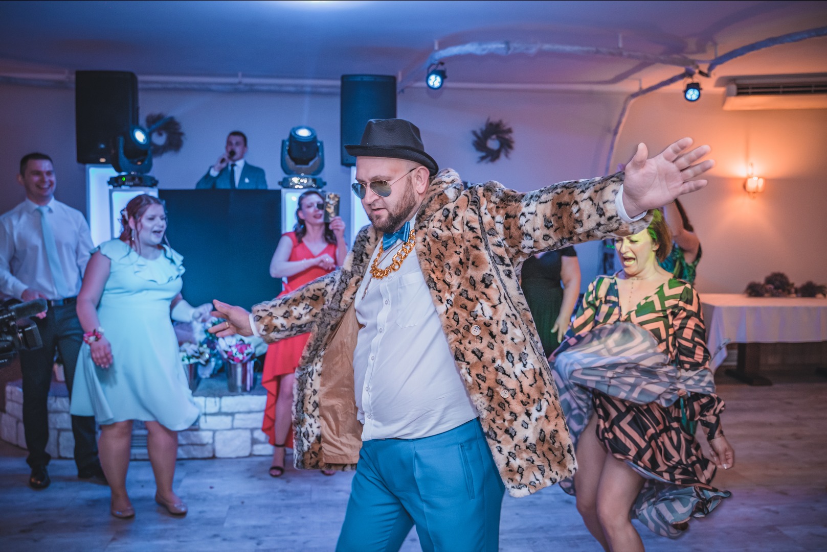 Mężczyzna w panterkowym płaszczu i kapeluszu tańczy na parkiecie podczas imprezy, w tle DJ i bawiący się goście.