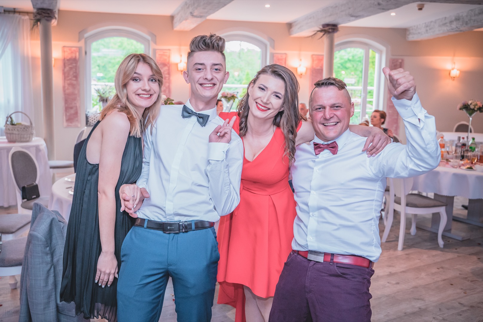 Grupa czterech uśmiechniętych osób pozuje do zdjęcia na sali weselnej, z widocznymi stołami i oknami w tle.