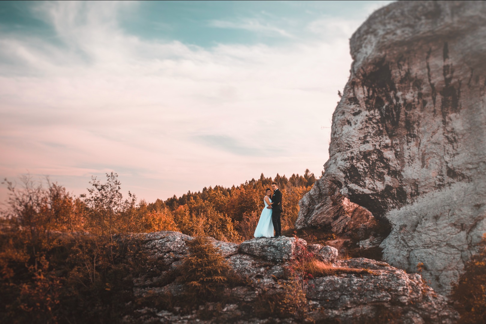 Para młoda w objęciach na skalistym wzgórzu, w tle jesienny las i formacja skalna z wyraźnym otworem. Ciepłe światło zachodzącego słońca.