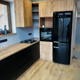 Nowoczesna kuchnia z frontami szafek w odcieniach drewna i czarnymi dolnymi szafkami, czarna lodówka SMEG, drewniany blat i płyta gazowa.