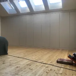 Wysoka zabudowa szafy wnękowej z jasnymi frontami, dopasowana do skosu poddasza, drewniana podłoga z widocznymi sękami, okna dachowe wpuszczające naturalne światło.