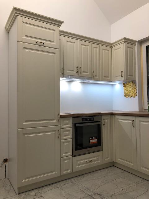 Jasna, klasyczna zabudowa kuchenna z frezowanymi frontami, widoczny piekarnik w zabudowie, drewniany blat i złote heksagonalne płytki na ścianie.