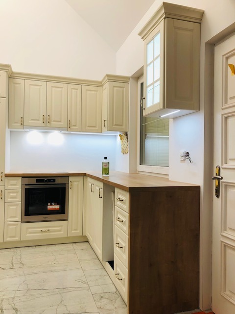 Jasna kuchnia w stylu klasycznym z drewnianym blatem, oświetleniem LED pod szafkami i marmurową podłogą. Widoczny piekarnik i butelka oliwy na blacie.