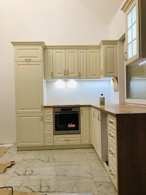 Kremowa zabudowa kuchenna w stylu klasycznym, z drewnianym blatem, piekarnikiem i oświetleniem podszafkowym, widoczna podczas montażu z zabezpieczeniem podłogi.