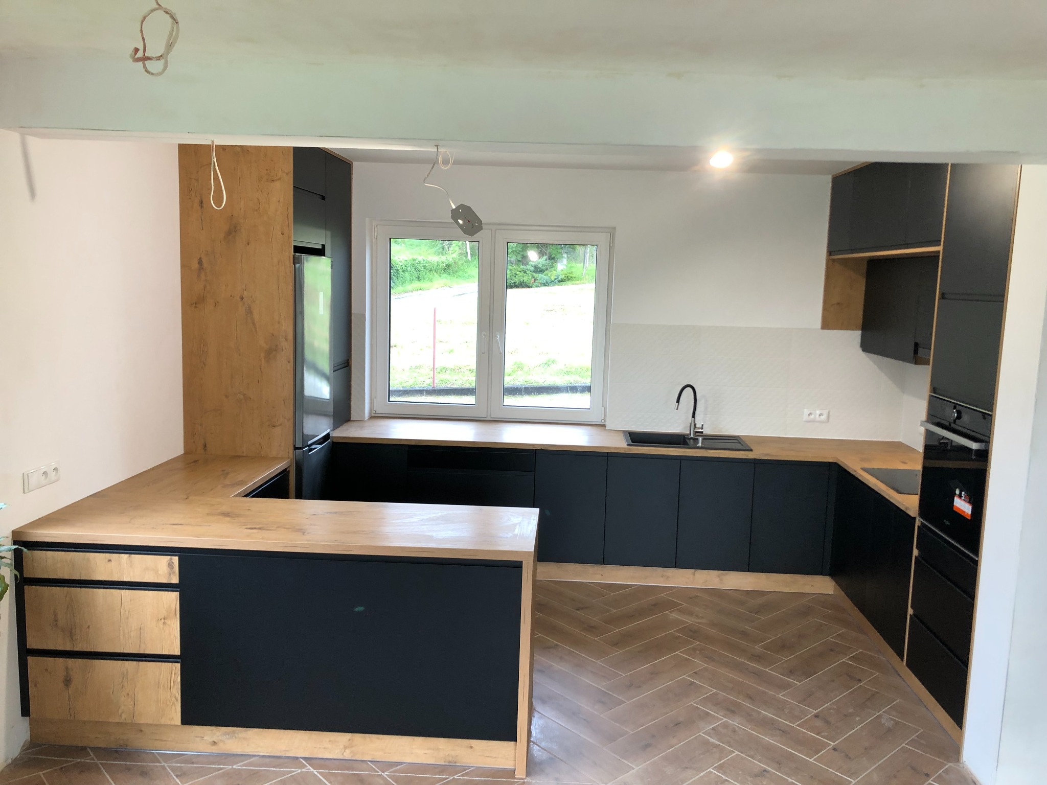 Nowoczesna kuchnia z czarnymi szafkami i drewnianymi blatami, widoczne niedokończone prace elektryczne przy suficie.