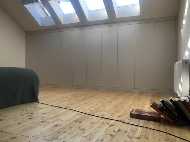 Wysoka zabudowa szafy wnękowej z jasnymi frontami, dopasowana do skosu poddasza, drewniana podłoga z widocznymi sękami, okna dachowe wpuszczające naturalne światło.