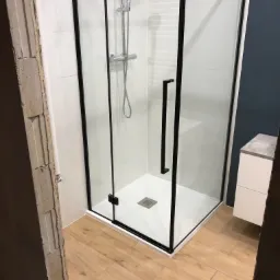 Nowoczesna kabina prysznicowa z czarnymi profilami, biały brodzik i armatura, widoczne białe płytki na ścianach i drewnopodobne panele podłogowe, fragment ściany w trakcie prac wykończeniowych po...