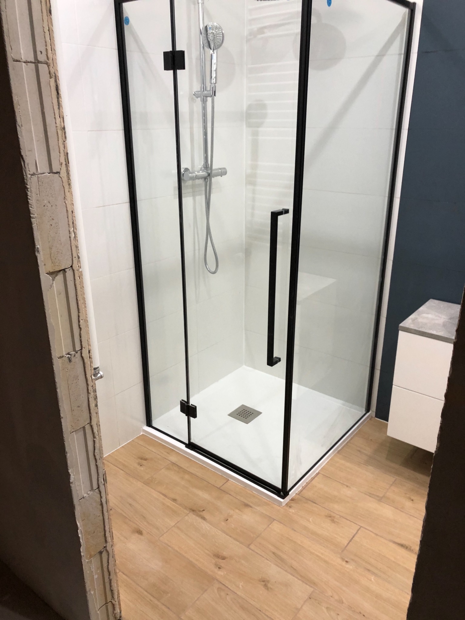 Nowoczesna kabina prysznicowa z czarnymi profilami, biały brodzik i armatura, widoczne białe płytki na ścianach i drewnopodobne panele podłogowe, fragment ściany w trakcie prac wykończeniowych po...