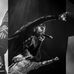 Czarno-białe zdjęcie rapera z brodą, w czapce z daszkiem i okularach przeciwsłonecznych, dynamicznie występującego z mikrofonem na scenie.
