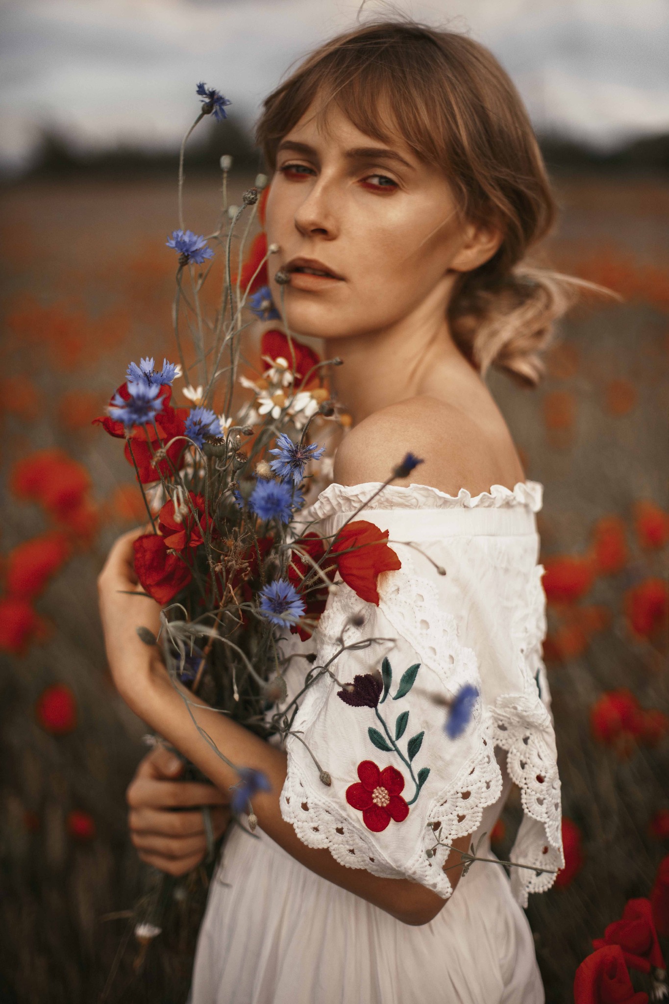 Kobieta w białej sukience z haftem kwiatowym trzyma bukiet polnych kwiatów na tle kwitnącego pola maków.