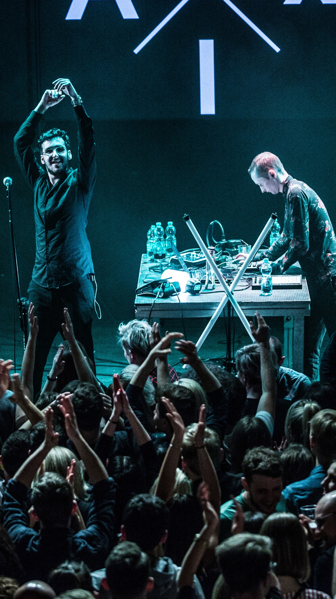 Entuzjastyczny DJ z podniesionymi rękami, grający muzykę elektroniczną na scenie podczas eventu, w tle drugi DJ przy sprzęcie i rozentuzjazmowany tłum.