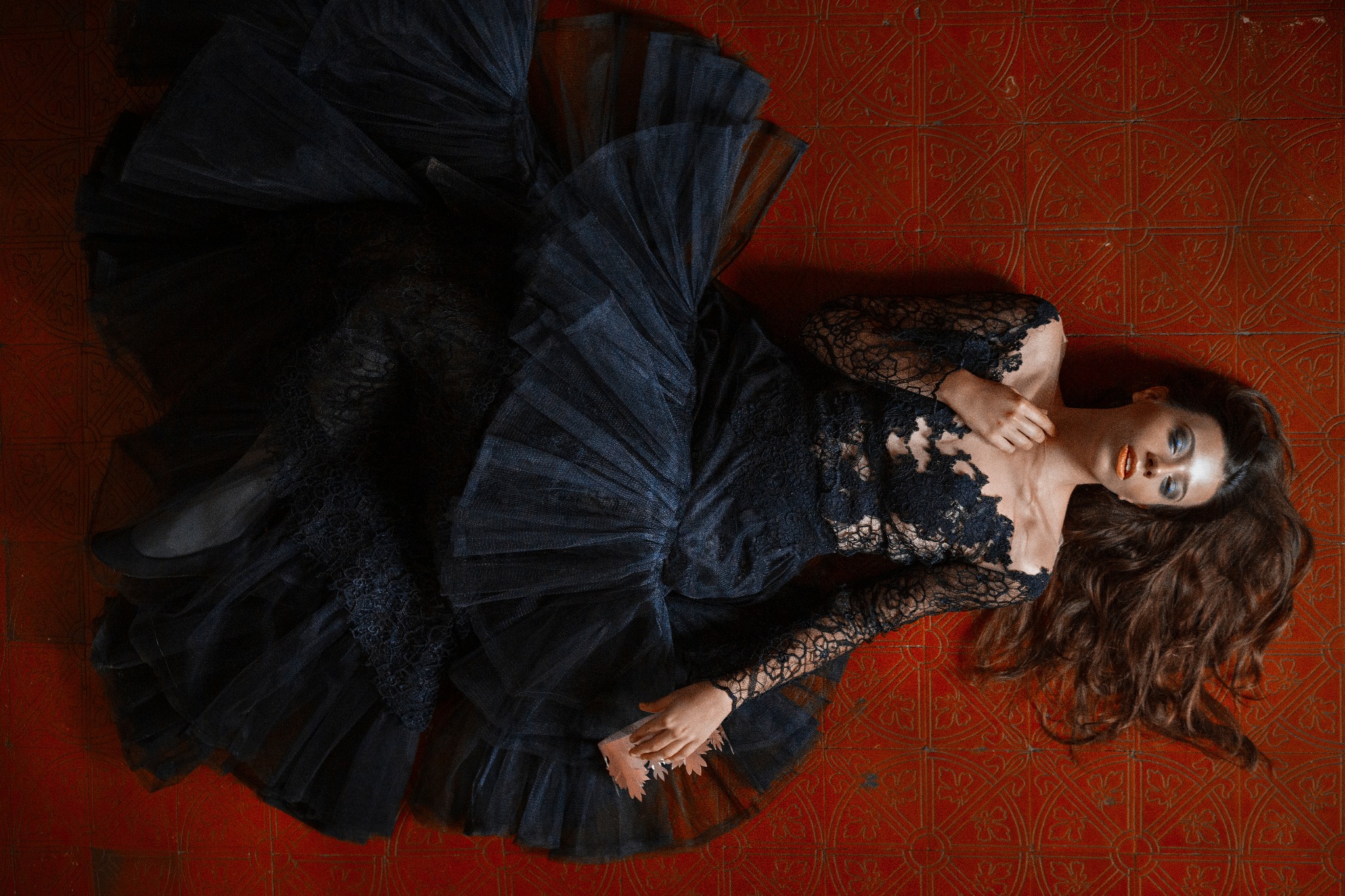 Kobieta leżąca na wzorzystej, czerwonej podłodze w czarnej, koronkowej sukni z tiulowym dołem, ułożona w artystycznej pozie, z rozpuszczonymi włosami i mocnym makijażem.