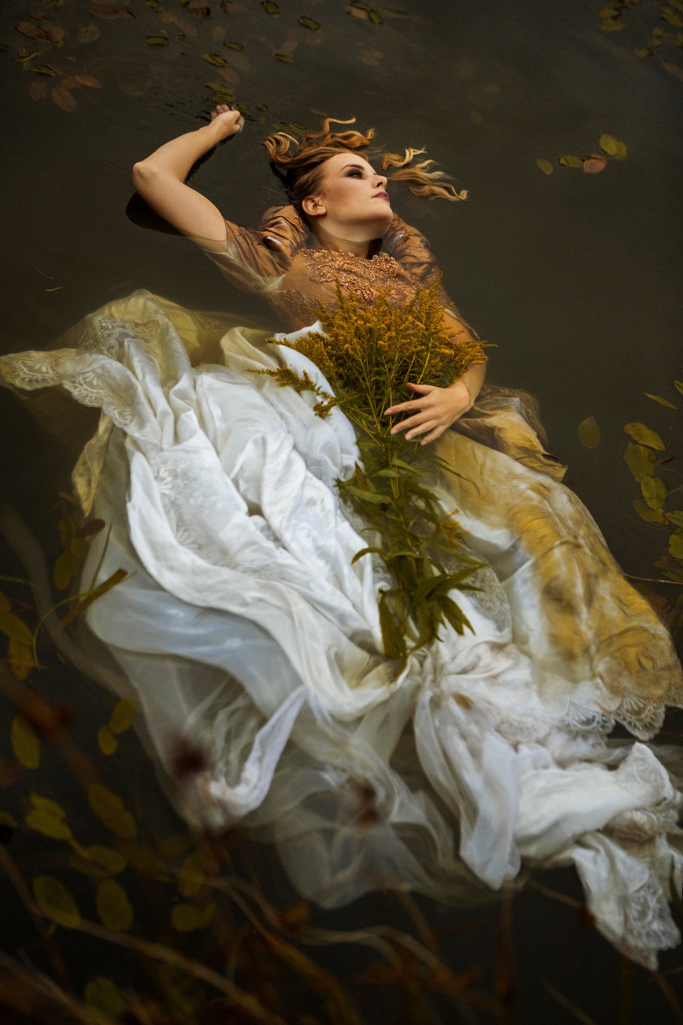 Kobieta w sukni unosi się na wodzie, trzymając bukiet żółtych kwiatów, z liśćmi na powierzchni wody.