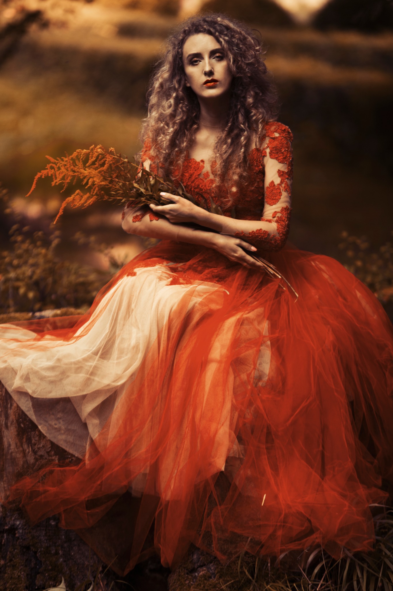 Kobieta w czerwono-kremowej sukni z tiulu i koronki, siedząca na kamieniu z bukietem rudych traw w zamyślonej pozie na tle leśnego krajobrazu w jesiennych barwach.
