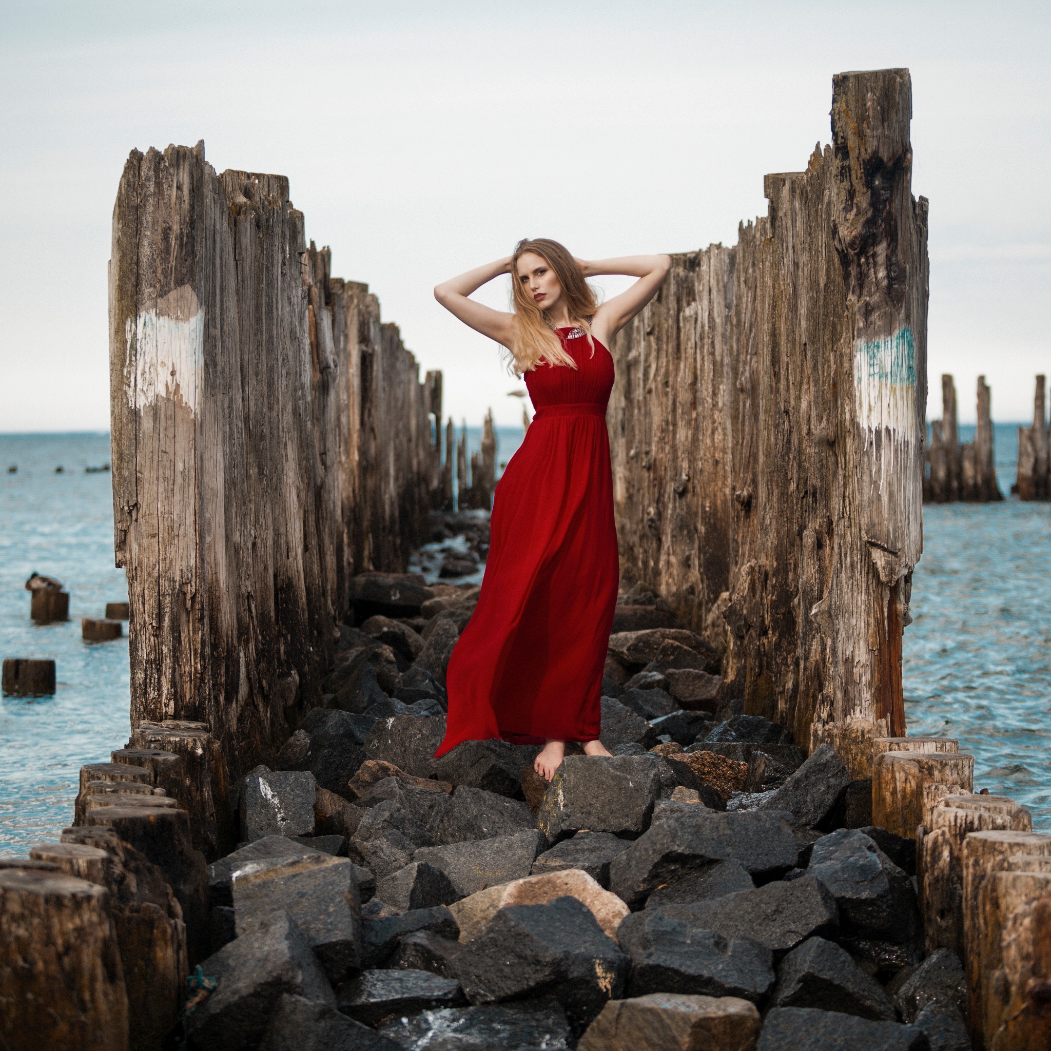 Kobieta w czerwonej sukni pozuje na tle morza, stojąc na kamieniach wśród drewnianych pali