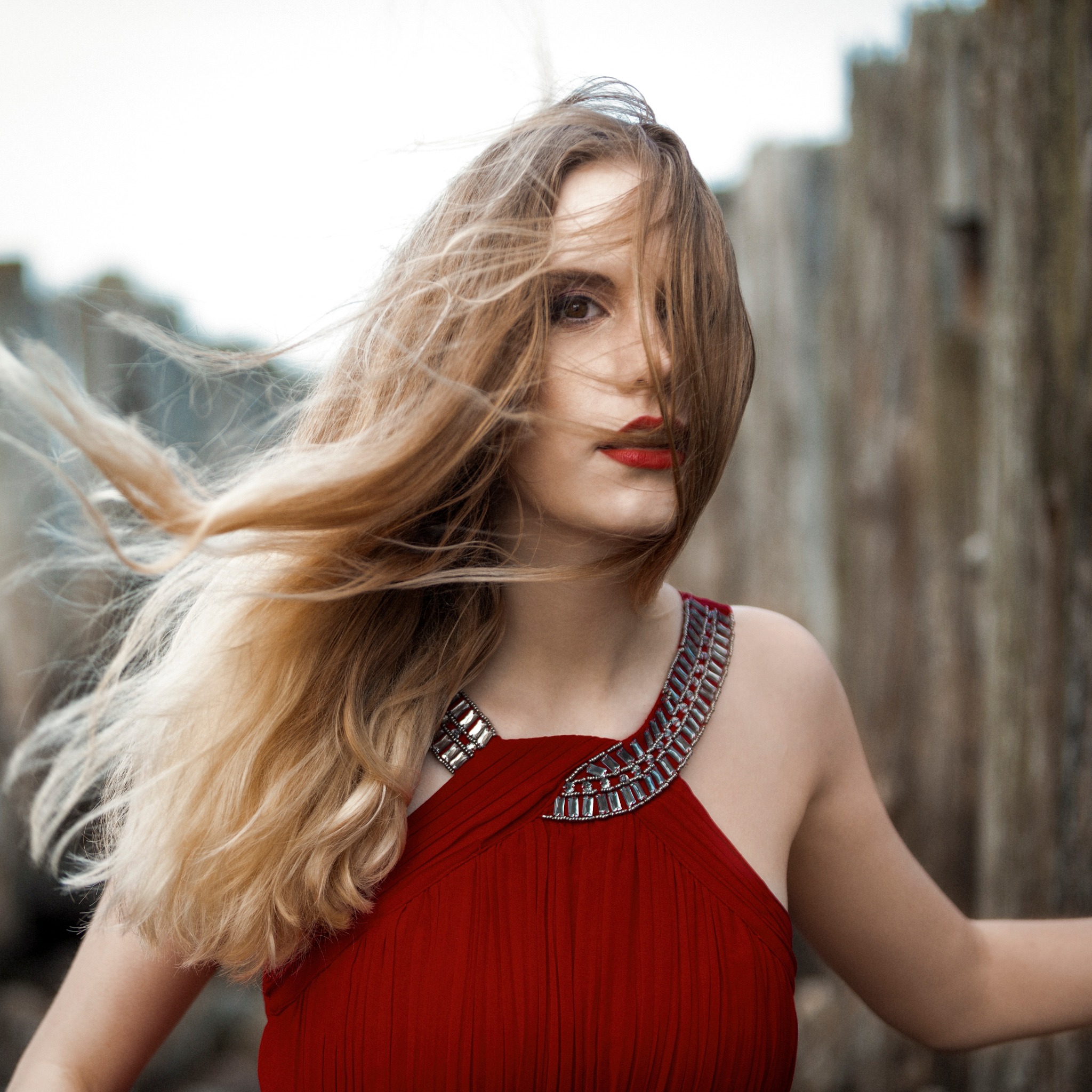 Portret kobiety w czerwonej sukience z ozdobnym dekoltem, włosy rozwiane na wietrze, na tle drewnianej konstrukcji.