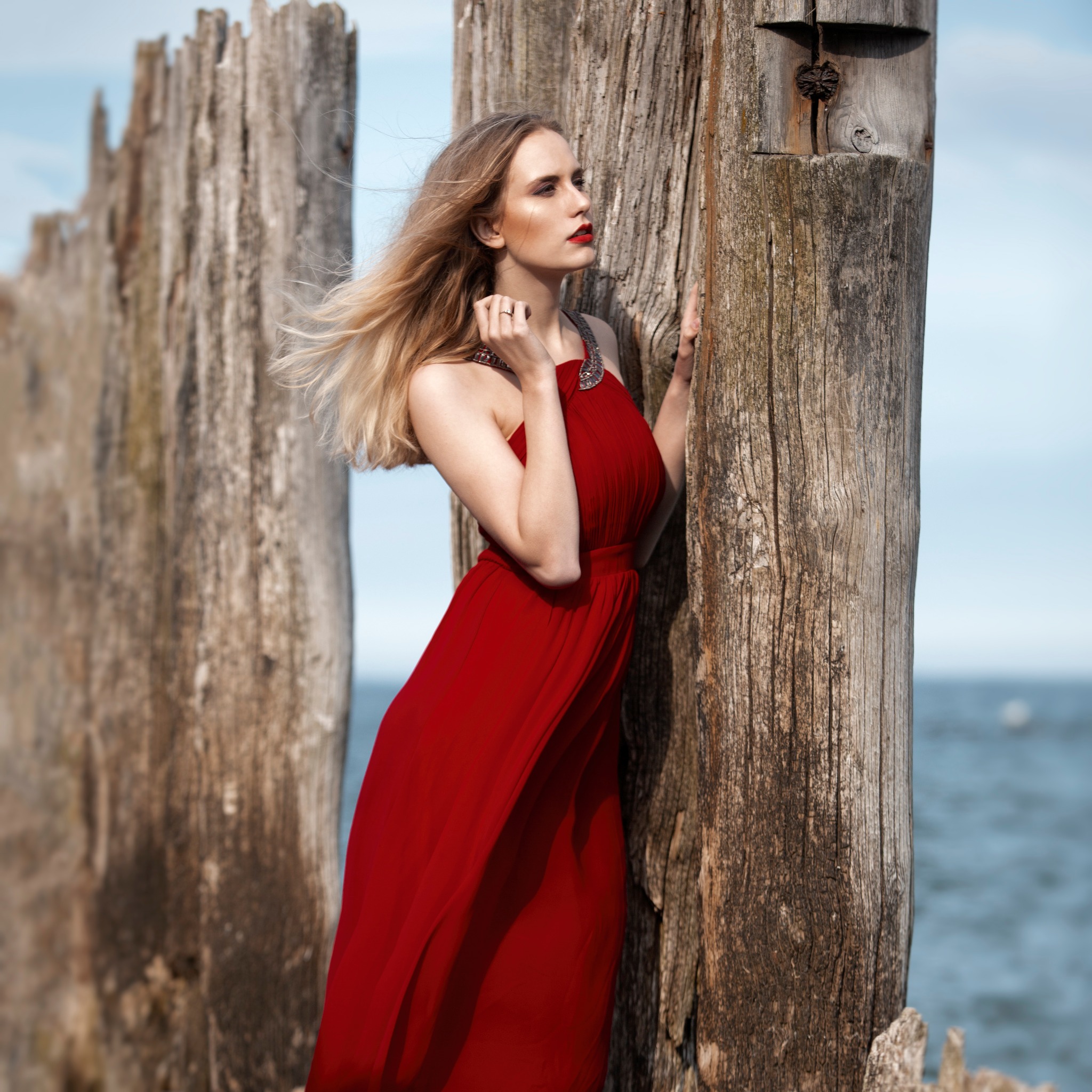 Blondwłosa kobieta w czerwonej sukni pozuje oparta o drewniane pale na tle morza, z wiatrem we włosach i mocnym makijażem.