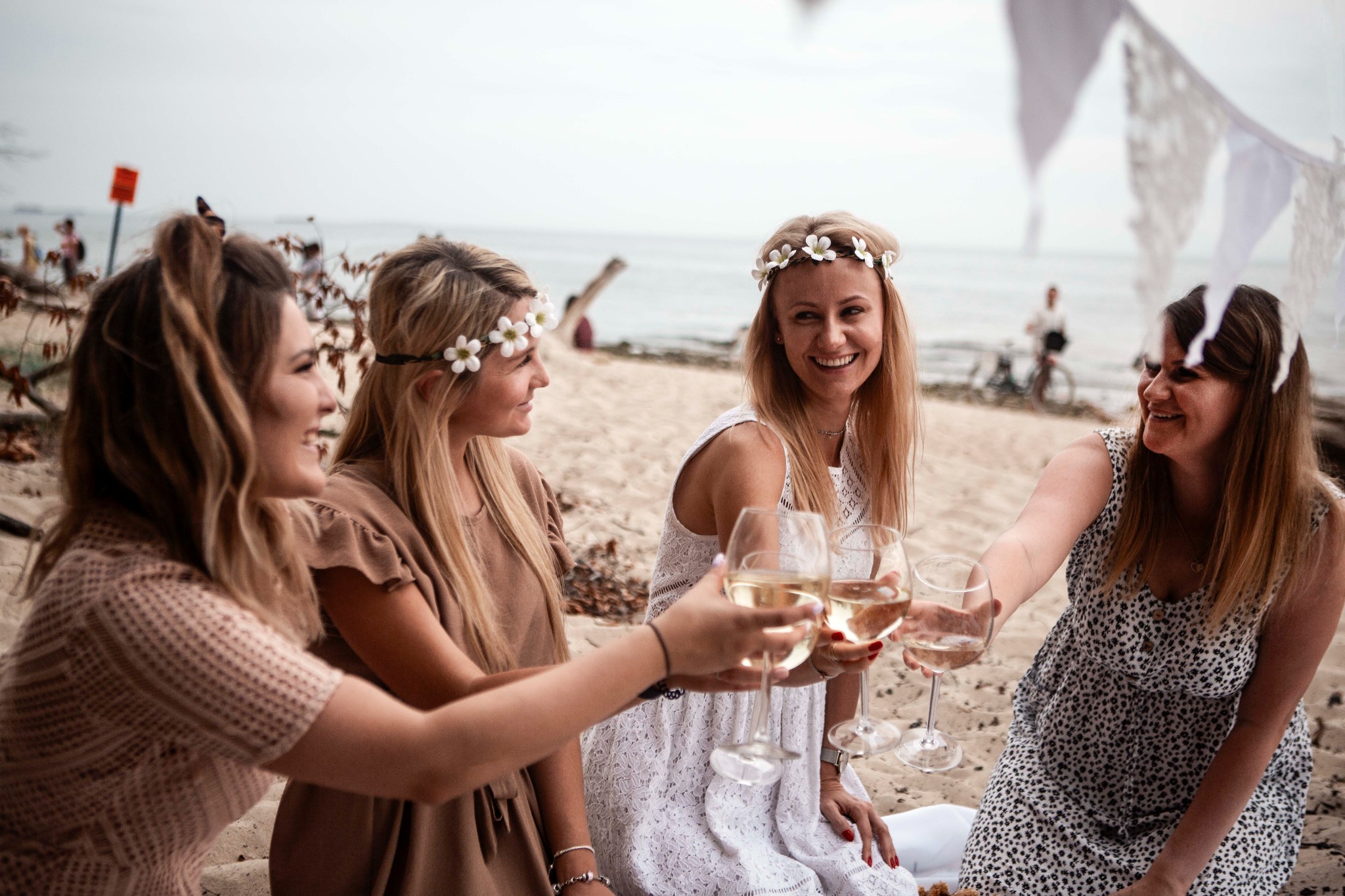 Cztery uśmiechnięte kobiety w kwiecistych wiankach na głowach, siedzące na piaszczystej plaży i wznoszące toast białym winem w kieliszkach, w tle dekoracje boho i sylwetka osoby na rowerze.