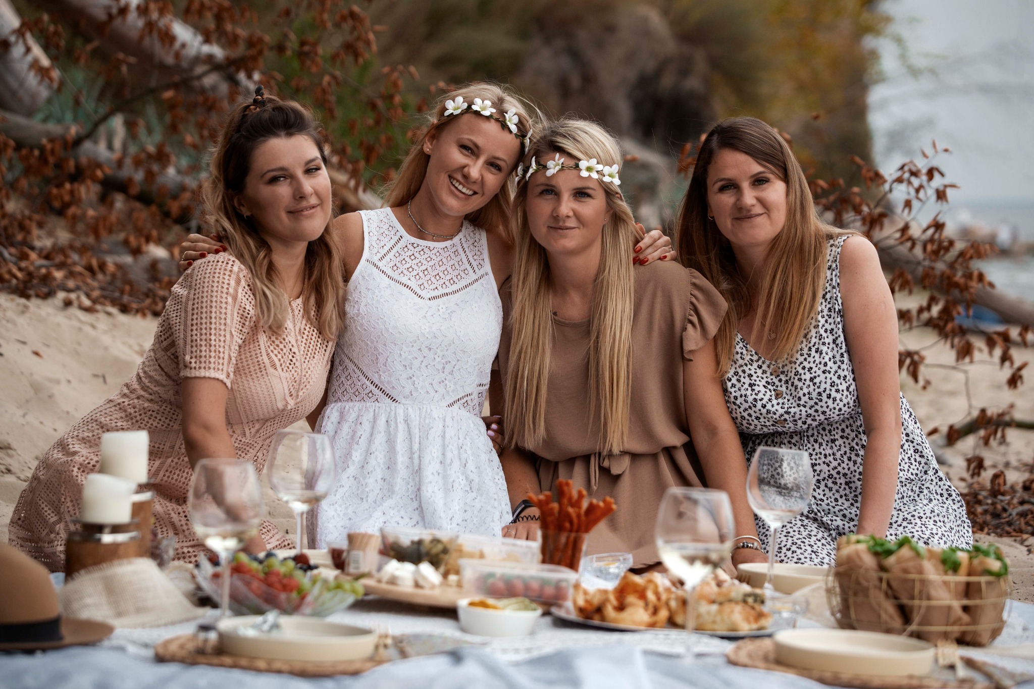 Cztery uśmiechnięte kobiety w kwiecistych wiankach na głowach, siedzące na plaży przy stole z przekąskami i winem, w tle widać drzewa i wodę.