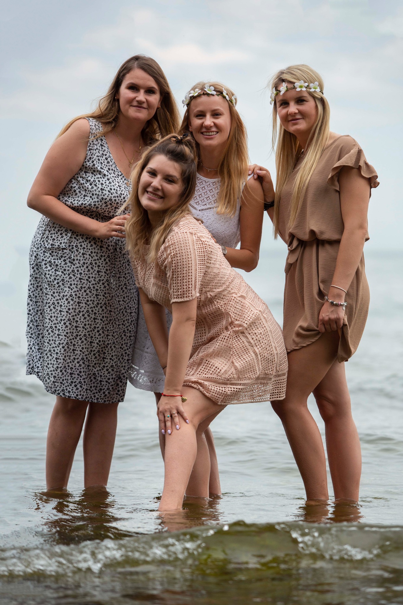 Cztery uśmiechnięte kobiety w letnich sukienkach i wiankach na głowach stoją w wodzie na brzegu morza, pozując do zdjęcia.