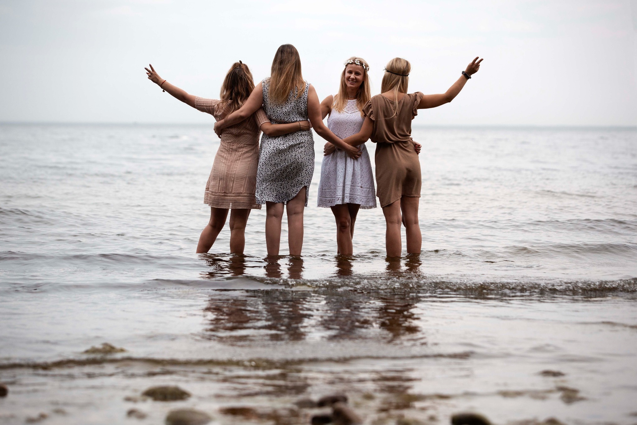 Cztery kobiety stojące w wodzie, obejmujące się, w letnich sukienkach, jedna z wiankiem na głowie, z podniesionymi rękami na tle morza.