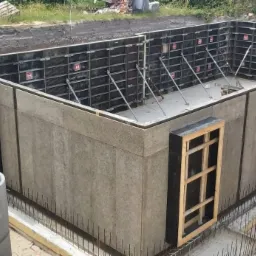 Szare szalunki z otworami okiennymi, zabezpieczone stalowymi prętami, na placu budowy z wykopem w tle.