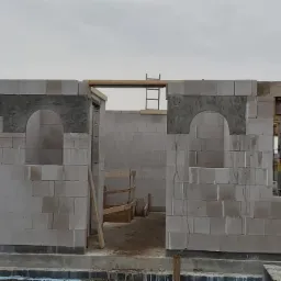 Wznoszenie ścian parterowego domu z betonu komórkowego, widoczne otwory okienne i drzwiowe, częściowo zamurowane łuki nad oknami, pracownik na rusztowaniu w tle.