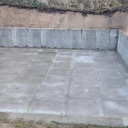 Henryk Jasiak Usługi Budowlane - Wykop z betonowymi ścianami oporowymi i wylewką z płyt betonowych na dnie, otoczony ziemią i trawą.