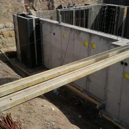 Henryk Jasiak Usługi Budowlane - Wykop pod fundamenty domu z wylanym betonem i zbrojeniem, widoczne drewniane stemple podtrzymujące szalunek, w tle budynki mieszkalne.