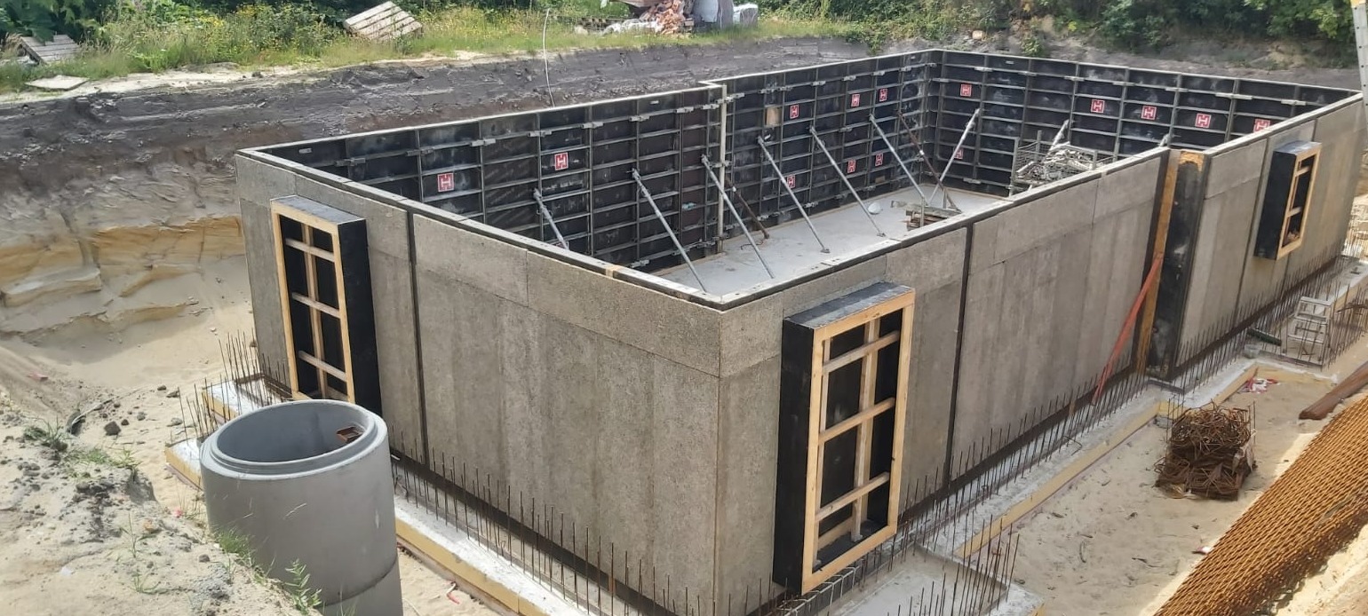 Szare szalunki z otworami okiennymi, zabezpieczone stalowymi prętami, na placu budowy z wykopem w tle.