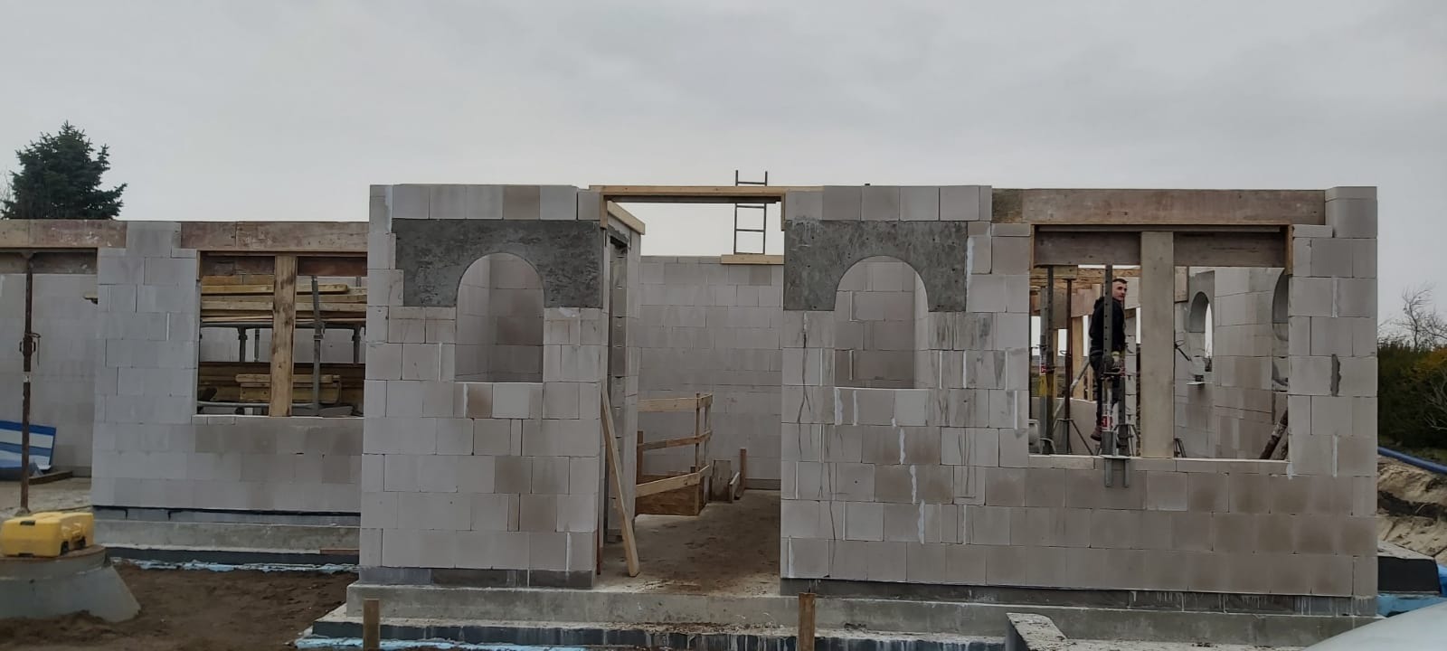 Wznoszenie ścian parterowego domu z betonu komórkowego, widoczne otwory okienne i drzwiowe, częściowo zamurowane łuki nad oknami, pracownik na rusztowaniu w tle.