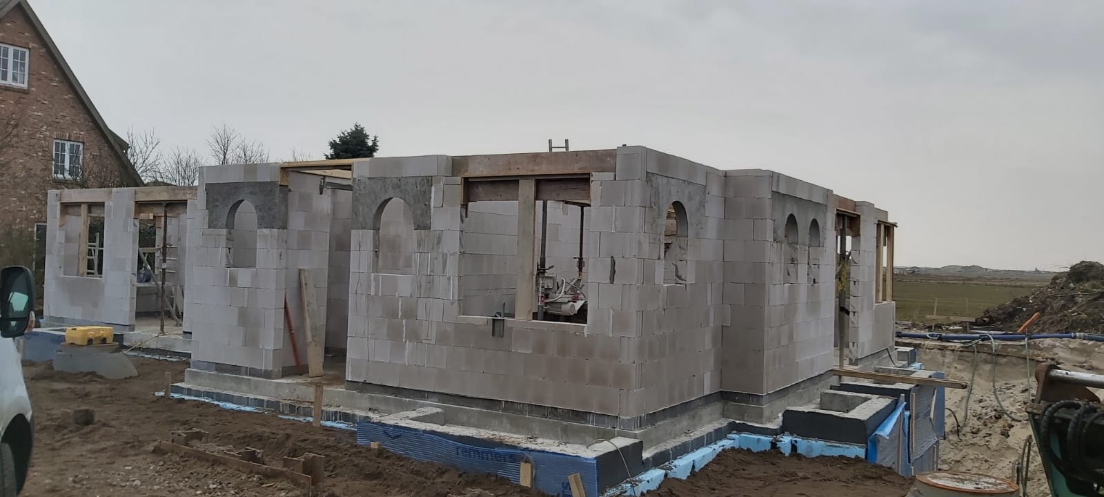 Wznoszenie ścian parterowego domu z bloczków silikatowych; widoczne otwory okienne z nadprożami łukowymi oraz izolacja fundamentów niebieskim styropianem.