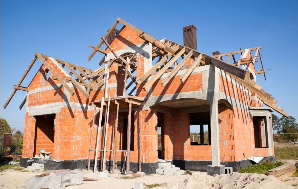 Surowy stan domu murowanego z czerwonej cegły, z drewnianą konstrukcją dachu w trakcie budowy, widoczny komin i fundamenty, na tle błękitnego nieba.