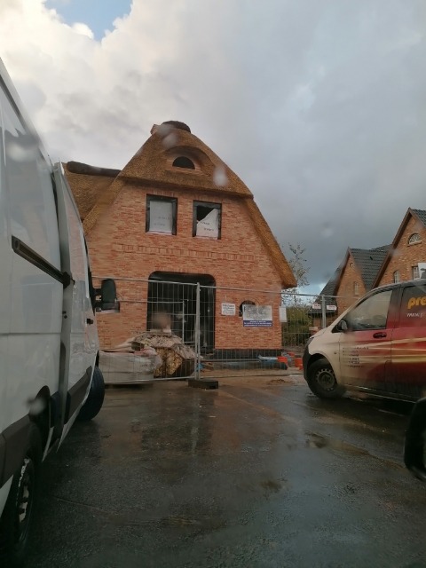 Budynek w trakcie budowy z cegły z dachem krytym strzechą, widoczne rusztowania i materiały budowlane, pochmurne niebo.